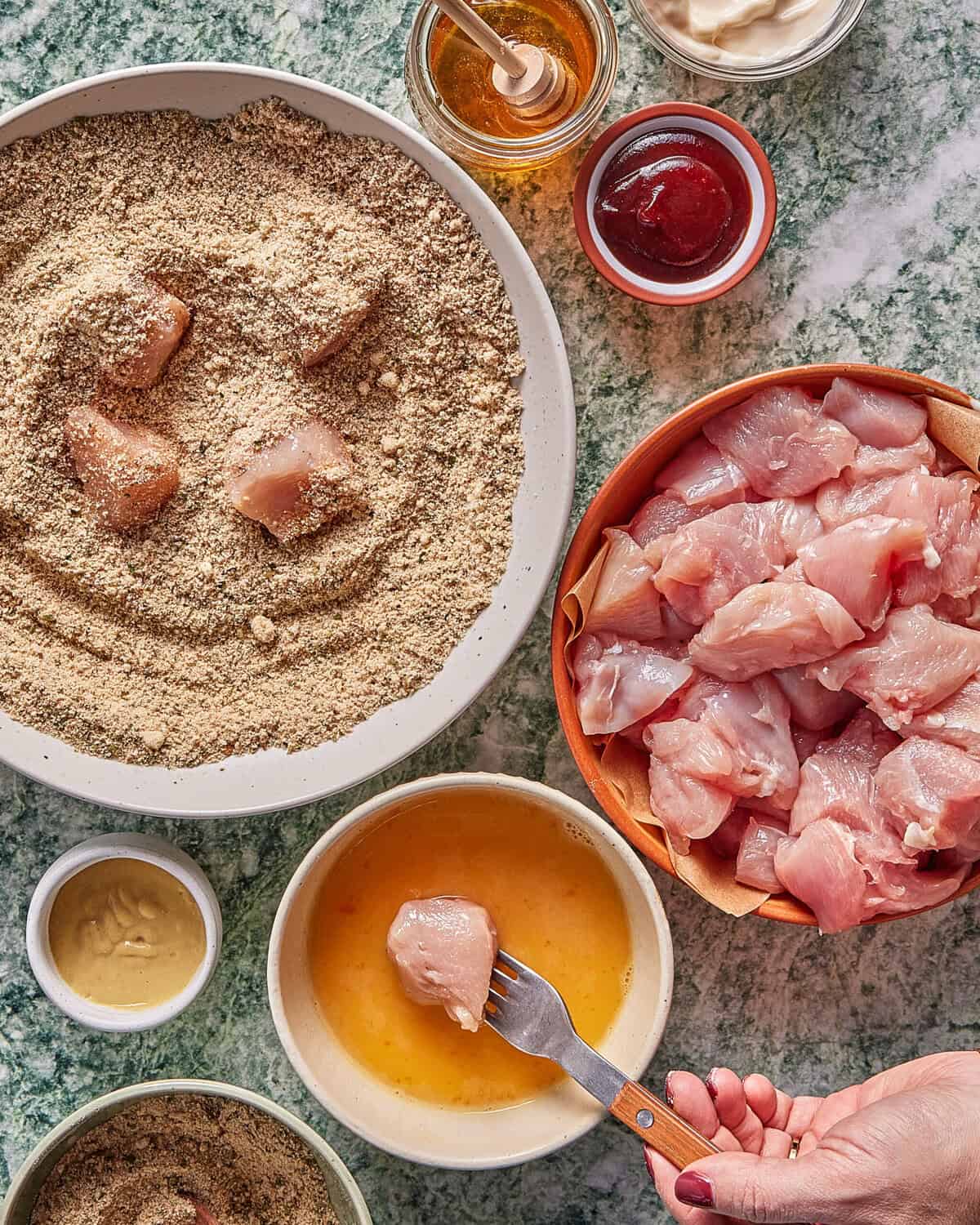 A hand dips a raw chicken piece into beaten egg. Nearby are bowls of raw chicken pieces, breadcrumbs, honey, ketchup, mayonnaise, and mustard, all on a green marbled countertop.
