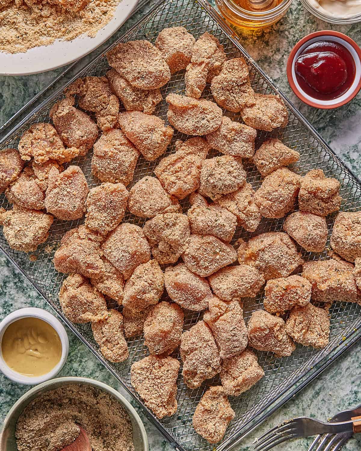 A tray filled with breaded chicken nuggets is surrounded by small bowls of dipping sauces and extra breading, all set on a marble countertop.