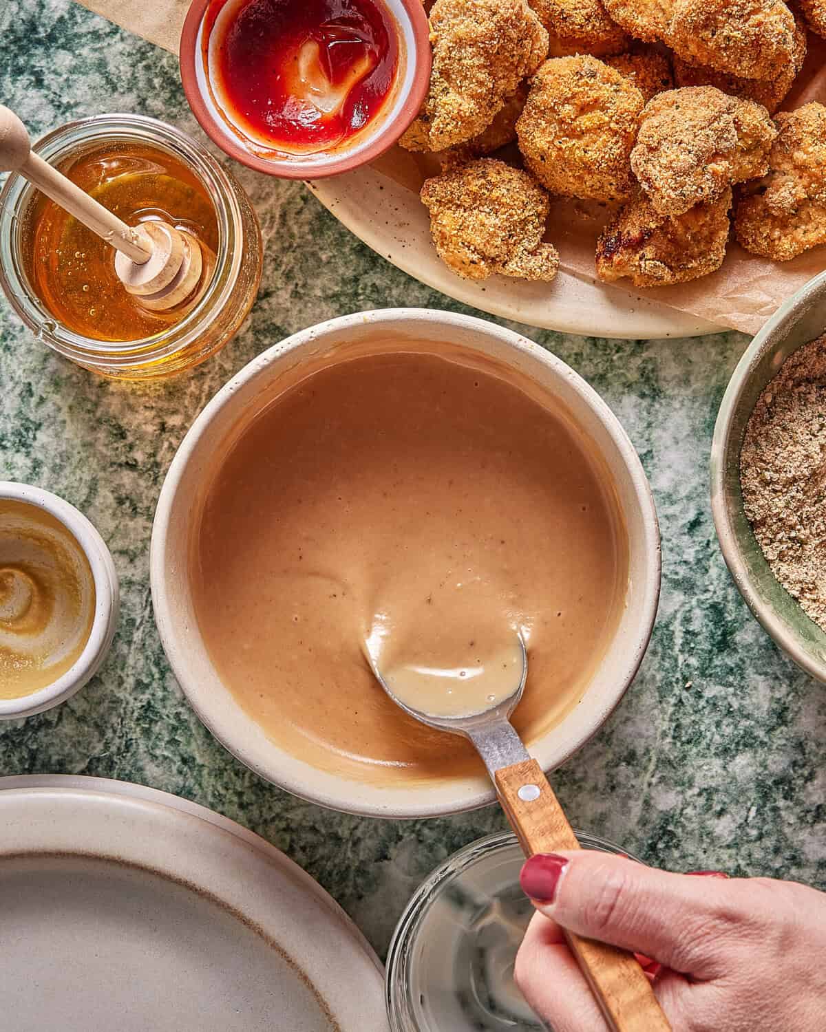 A hand holds a spoon over a bowl of creamy dipping sauce, surrounded by honey, mustard, ketchup, breaded nuggets, and breadcrumbs on a green marble surface.