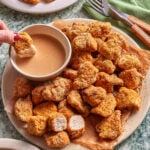 A plate of crispy, breaded chicken nuggets on parchment paper, with one nugget being dipped into a bowl of creamy sauce. A fork and knife rest on a green napkin nearby.
