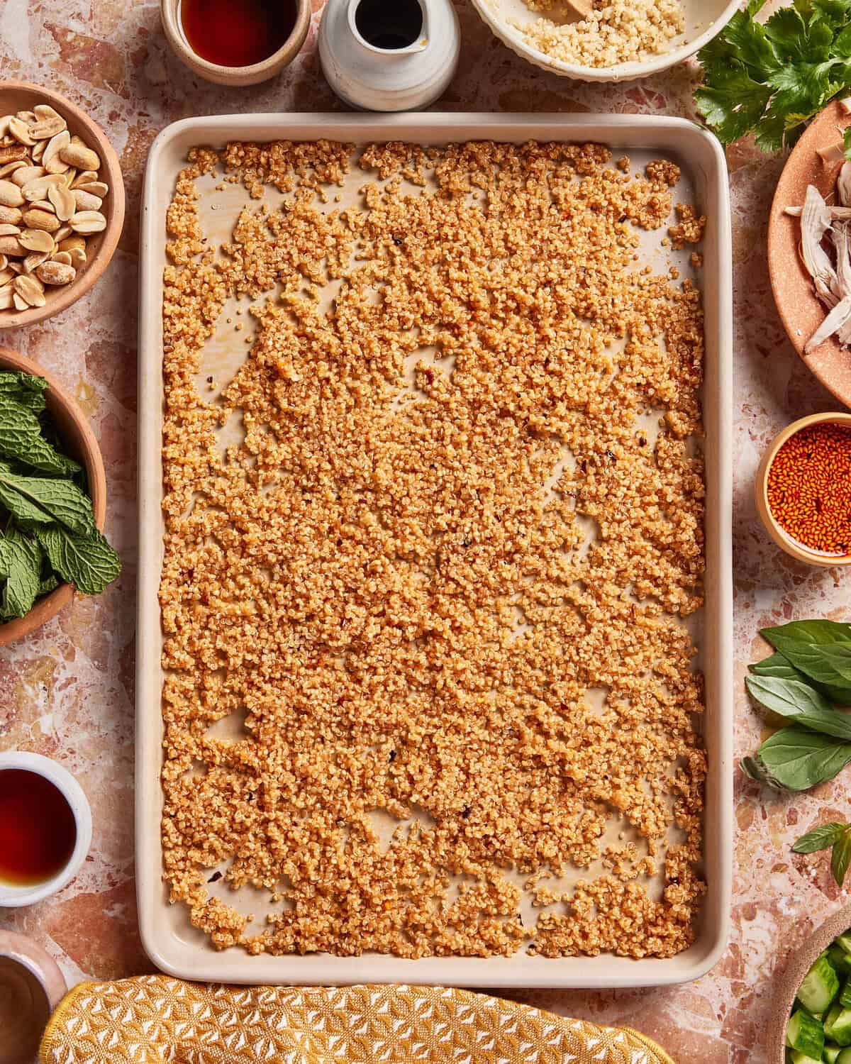 A baking tray of cooked, fluffed couscous sits on a counter surrounded by small bowls of herbs, nuts, sauces, and other ingredients, ready for meal preparation.