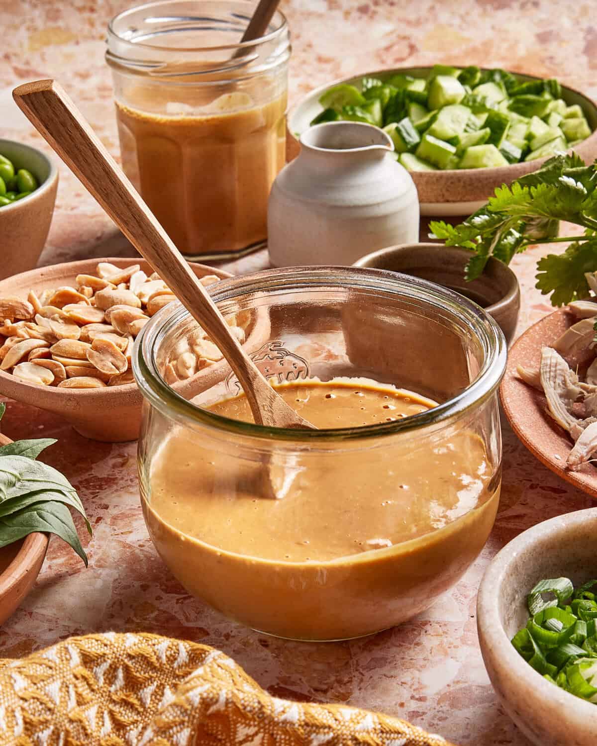 A glass jar filled with creamy peanut sauce sits on a counter surrounded by bowls of peanuts, cucumbers, herbs, and a small pitcher, with a wooden spoon resting in the jar.