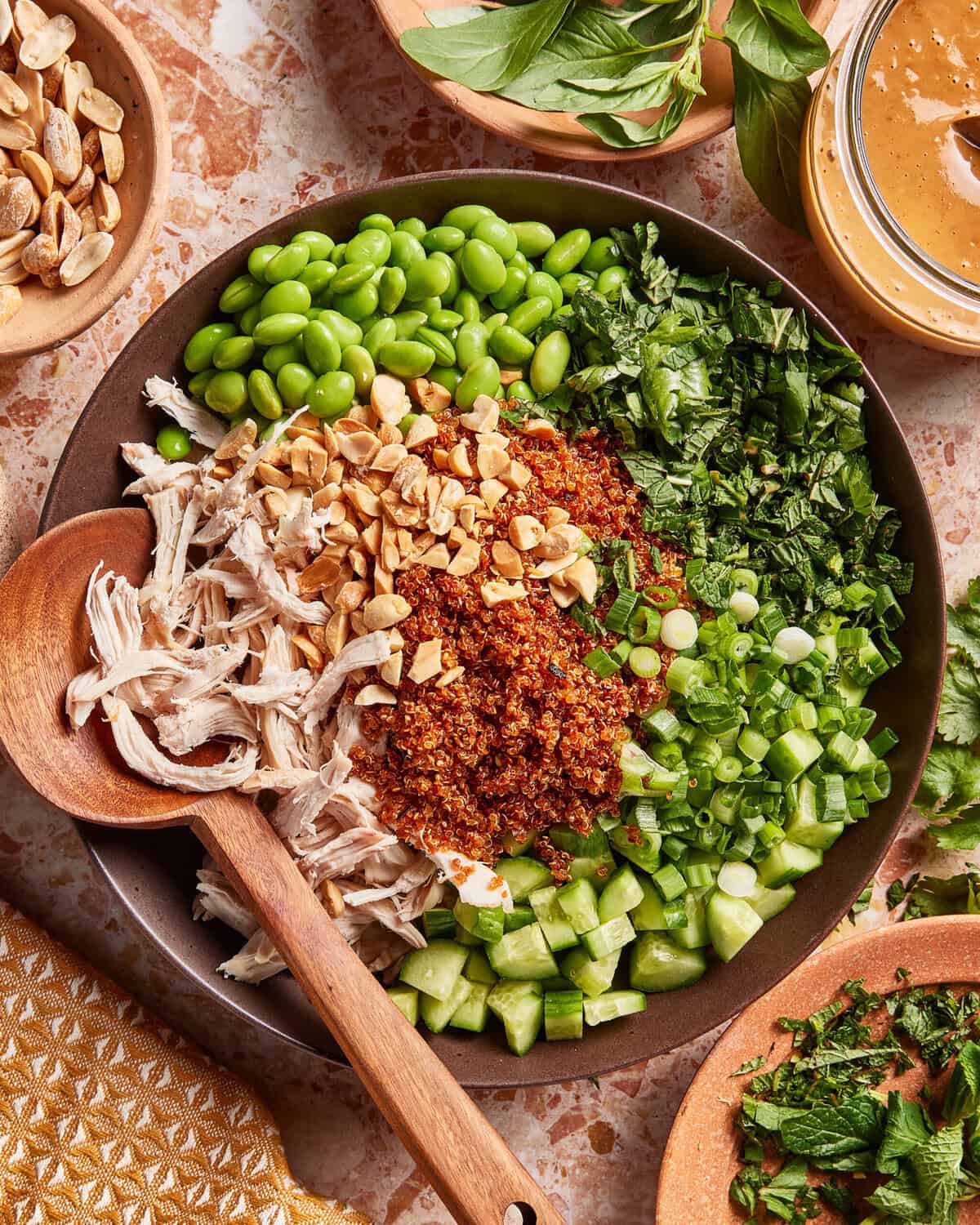 A bowl filled with shredded chicken, diced cucumber, chopped herbs, edamame, green onions, crushed peanuts, and chili sauce, with a wooden spoon on the side and fresh herbs and sauce nearby.