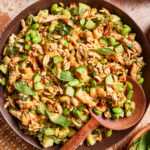 A bowl of salad with shredded chicken, cucumbers, edamame, peanuts, fresh herbs, and a creamy tan dressing, served with a wooden spoon on a patterned surface.