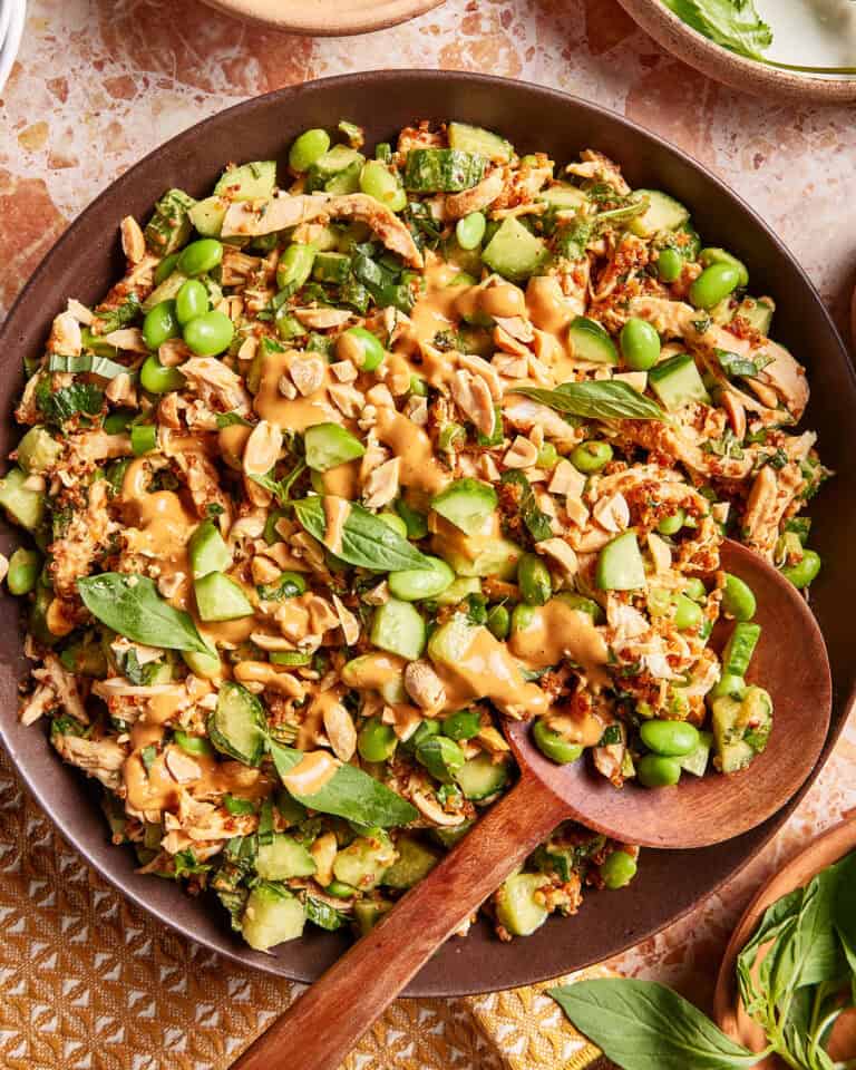 A bowl of salad with shredded chicken, cucumbers, edamame, peanuts, fresh herbs, and a creamy tan dressing, served with a wooden spoon on a patterned surface.