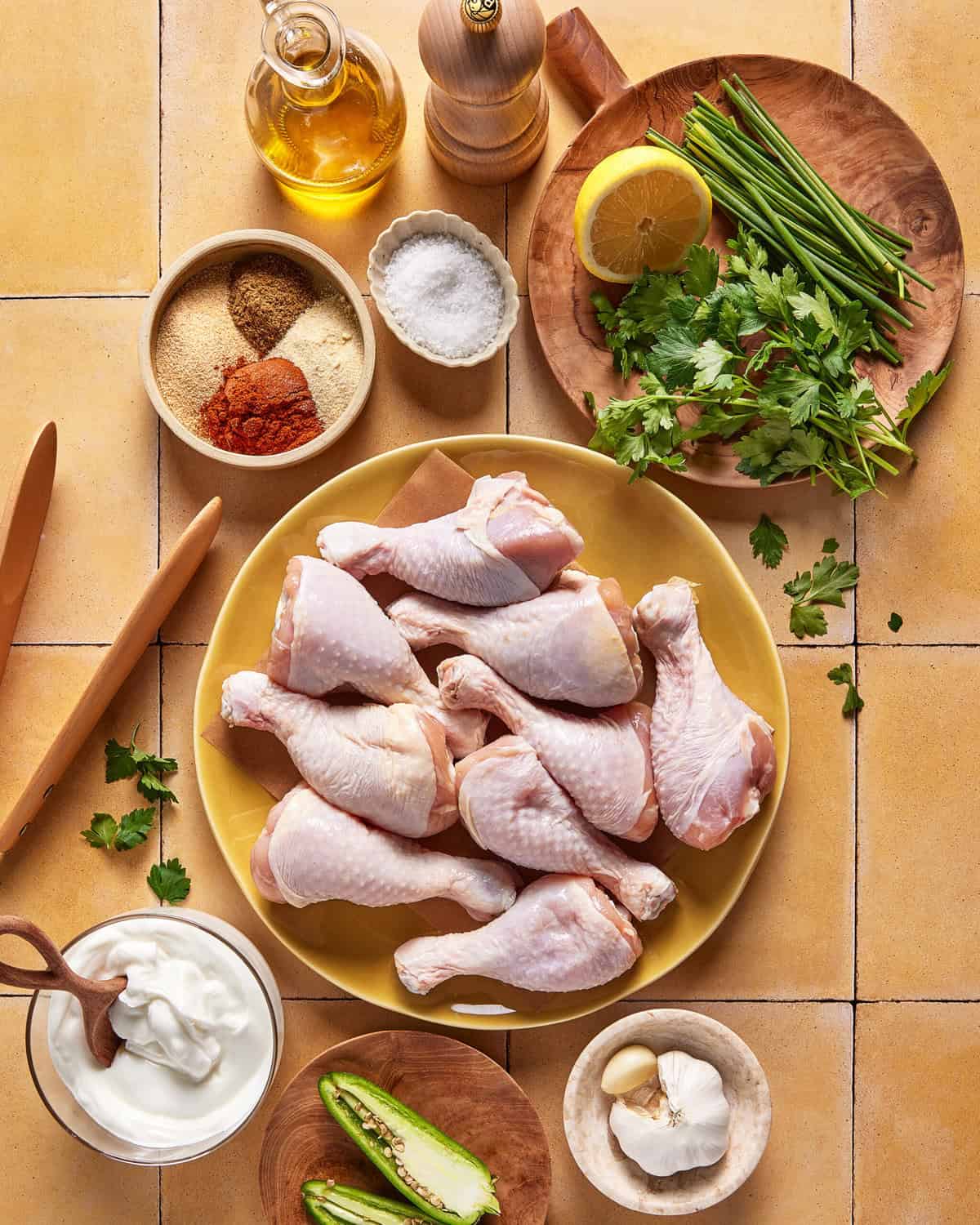 A yellow plate of raw chicken drumsticks is surrounded by small bowls of spices, salt, yogurt, fresh herbs, a halved jalapeño, garlic, a lemon half, wooden utensils, and a bottle of olive oil on a tiled countertop.