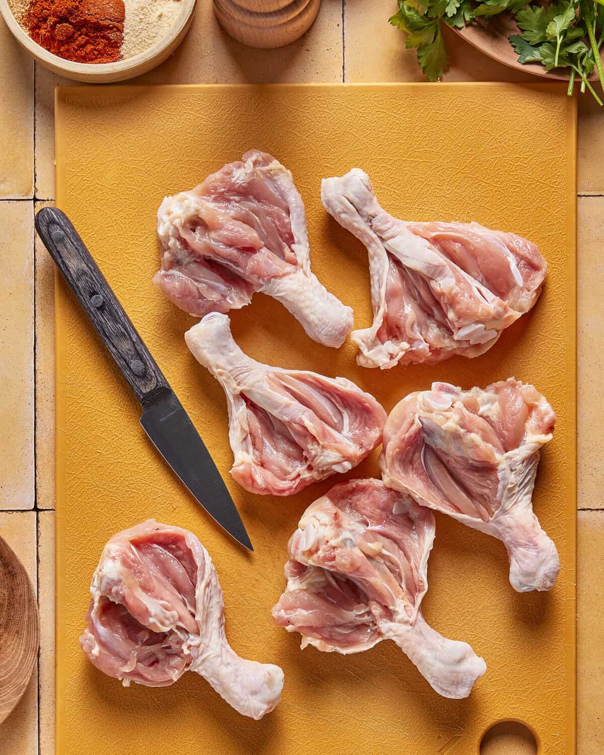 Six raw chicken drumsticks are arranged on a yellow cutting board with a small black-handled knife beside them. Some herbs and spices are partially visible around the edges of the board.