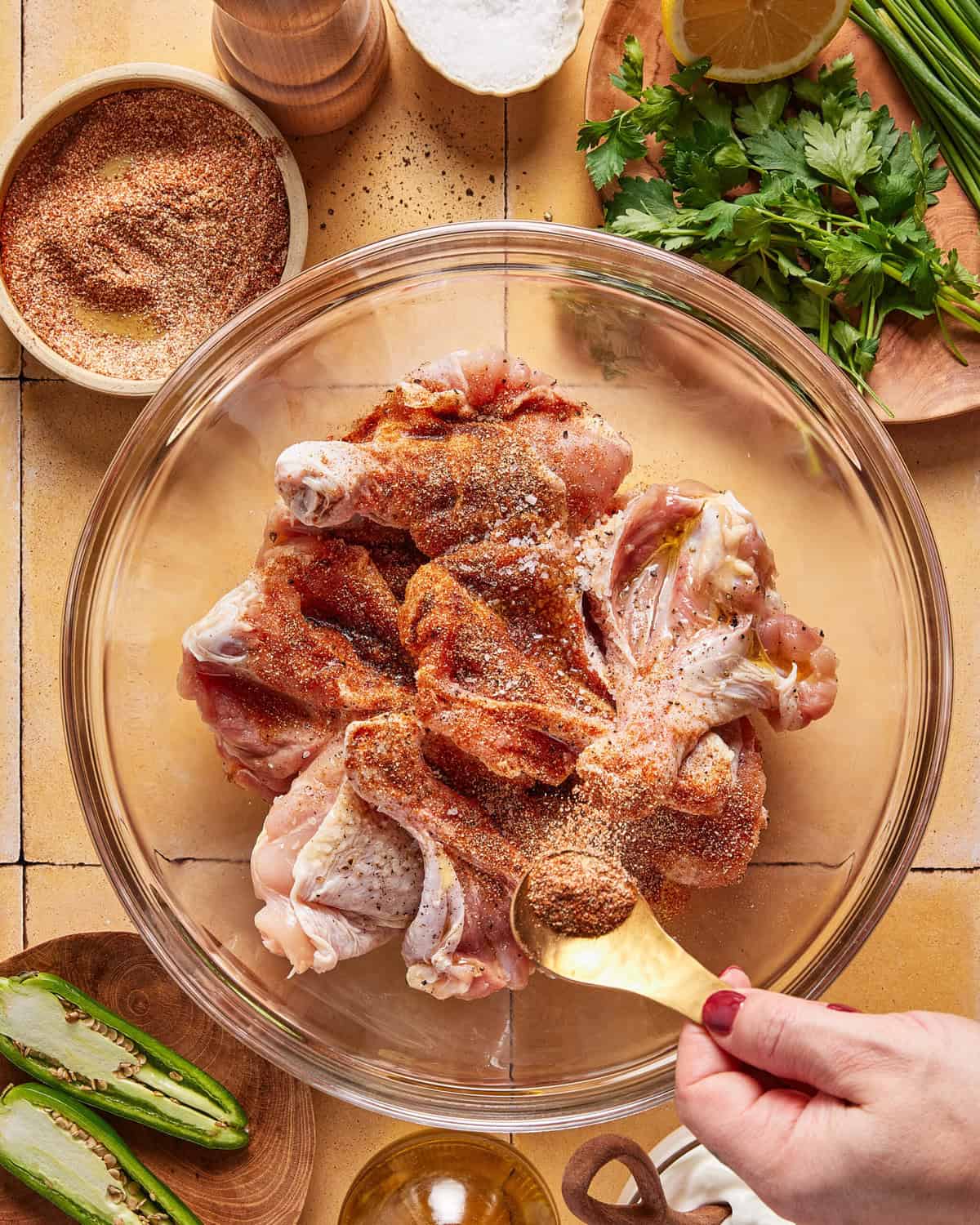 A hand sprinkles spice mixture onto raw chicken drumsticks in a glass bowl. Surrounding the bowl are fresh herbs, sliced jalapeños, lemon, a bowl of spices, and other cooking ingredients on a tiled surface.