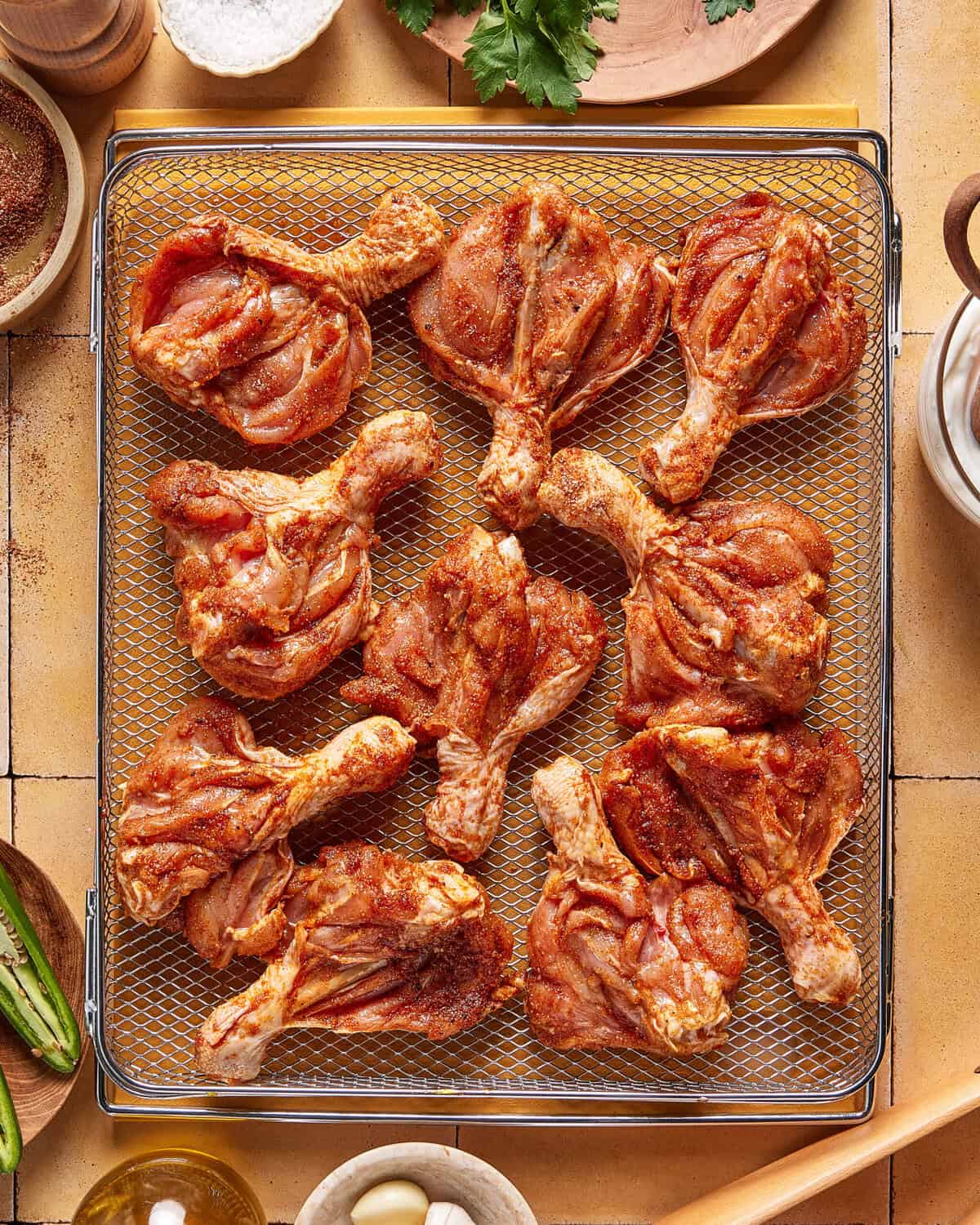 Raw, seasoned chicken drumsticks arranged in a single layer on a metal wire rack, ready for cooking. Surrounding the rack are bowls with spices, garlic, and herbs on a tiled countertop.