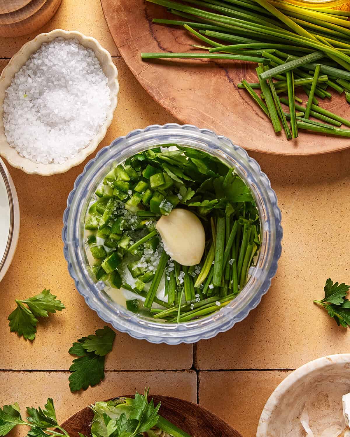 A food processor bowl containing chopped green onions, a garlic clove, diced green pepper, and coarse salt, surrounded by more fresh herbs and a dish of salt on a tiled surface.