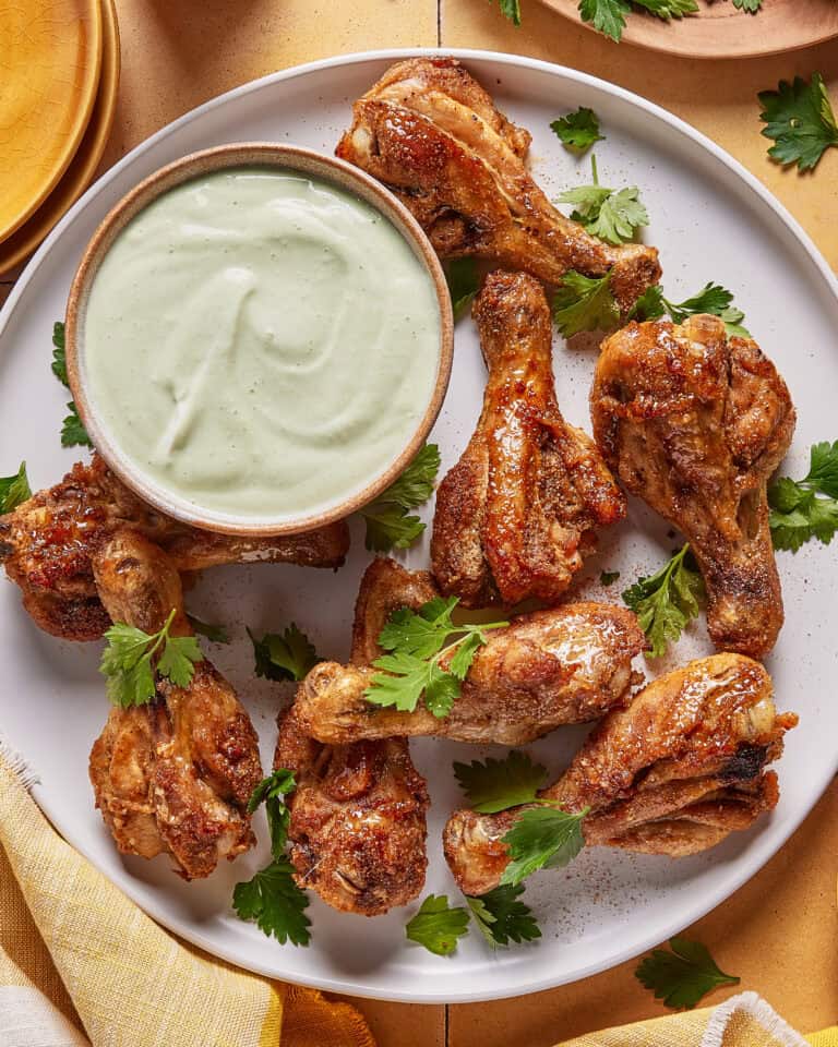 A white plate with roasted chicken drumsticks garnished with fresh parsley, served alongside a bowl of creamy dipping sauce.