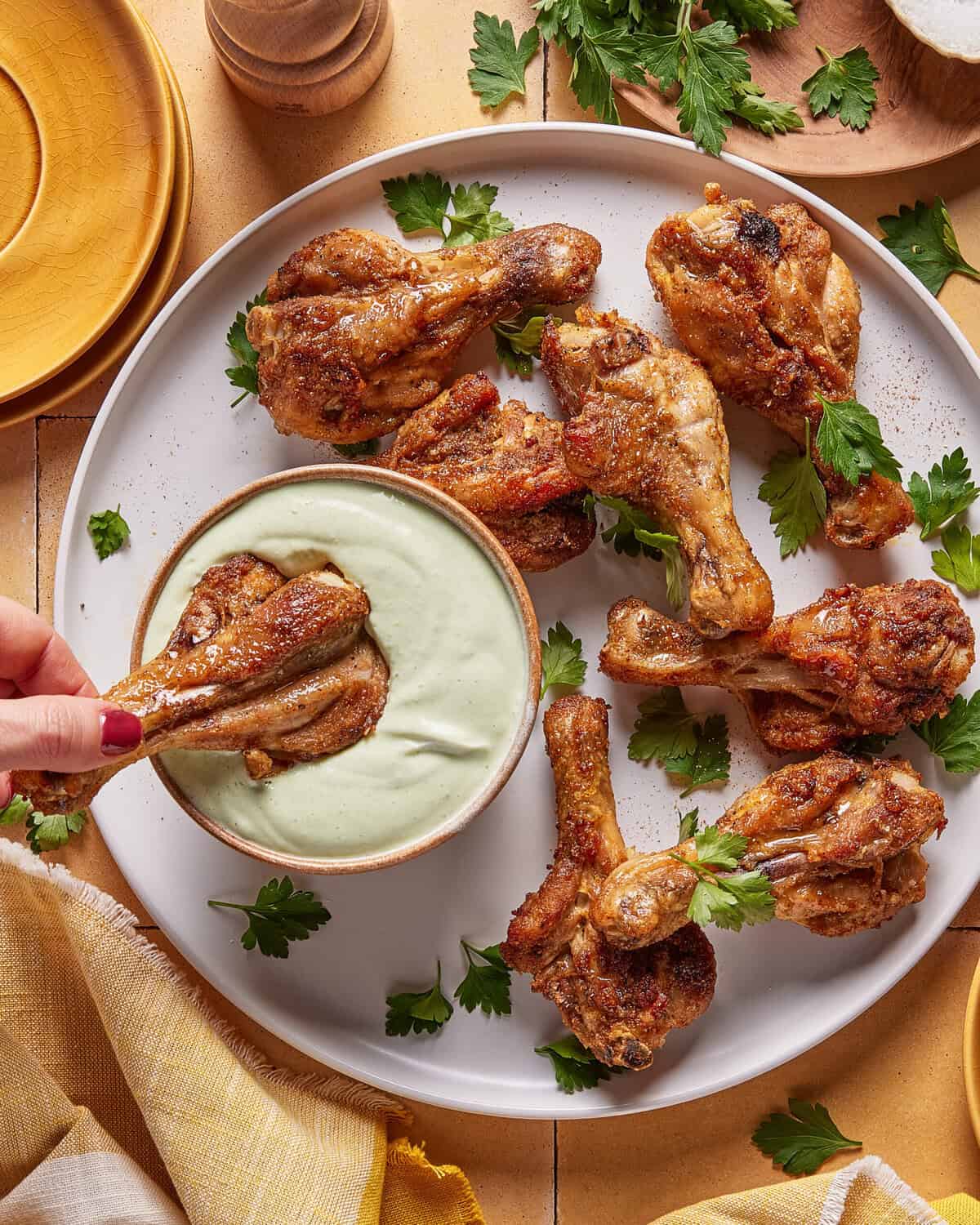 A plate of roasted chicken drumsticks garnished with parsley, with one drumstick being dipped into a bowl of creamy green sauce. Plates, a napkin, and herb sprigs surround the dish.