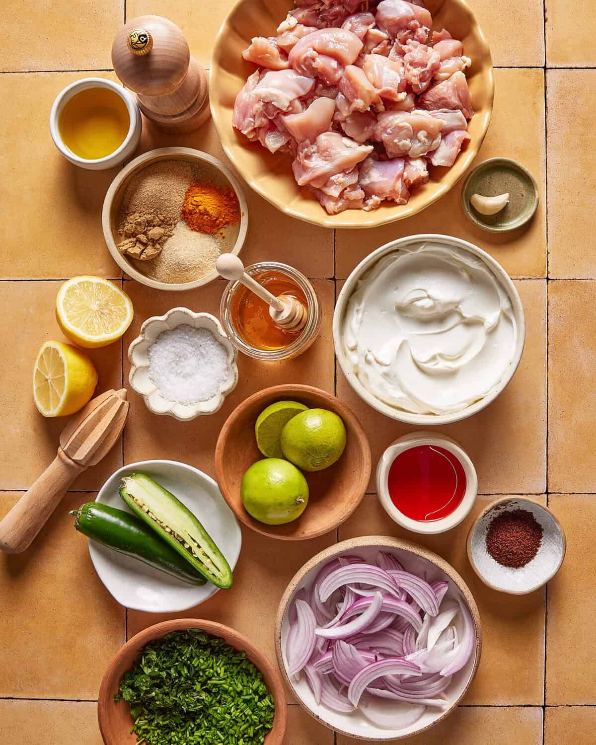 Various ingredients for cooking are arranged on a tiled surface, including raw chicken pieces, sliced onions, chopped cilantro, spices, yogurt, olive oil, lemon, lime, honey, a green chili, and a garlic clove.