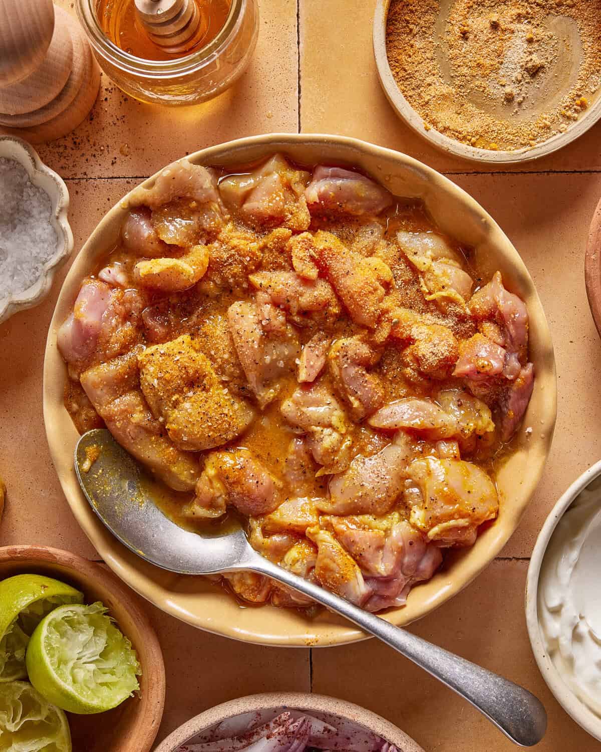 A bowl of marinated raw chicken pieces with spices, surrounded by a spoon, squeezed lime halves, salt, a jar of honey, spice powder, and a small bowl of yogurt on a tiled surface.