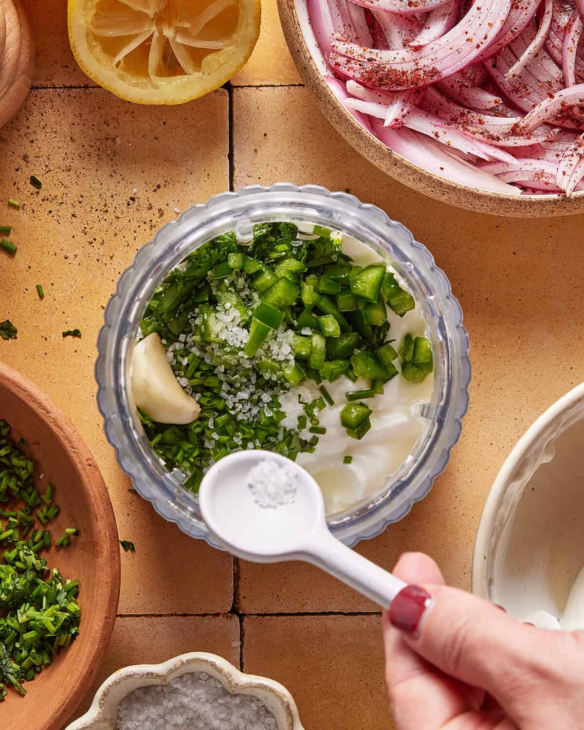 A hand holds a white spoon of salt over a jar filled with chopped green herbs, garlic, salt, and yogurt. Surrounding the jar are lemon, sliced red onions, and more chopped herbs in bowls.