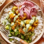 A bowl of white rice topped with diced cucumber, marinated chicken pieces in sauce, pickled red onions, and a drizzle of green creamy dressing, with a fork resting on the bowl’s edge.