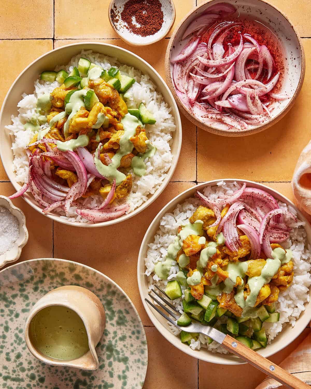Two bowls of rice topped with chopped cucumber, marinated chicken, sliced red onions, and green sauce, with extra onions and sauce on the side. The scene is set on a tiled surface with a fork and a ceramic plate.