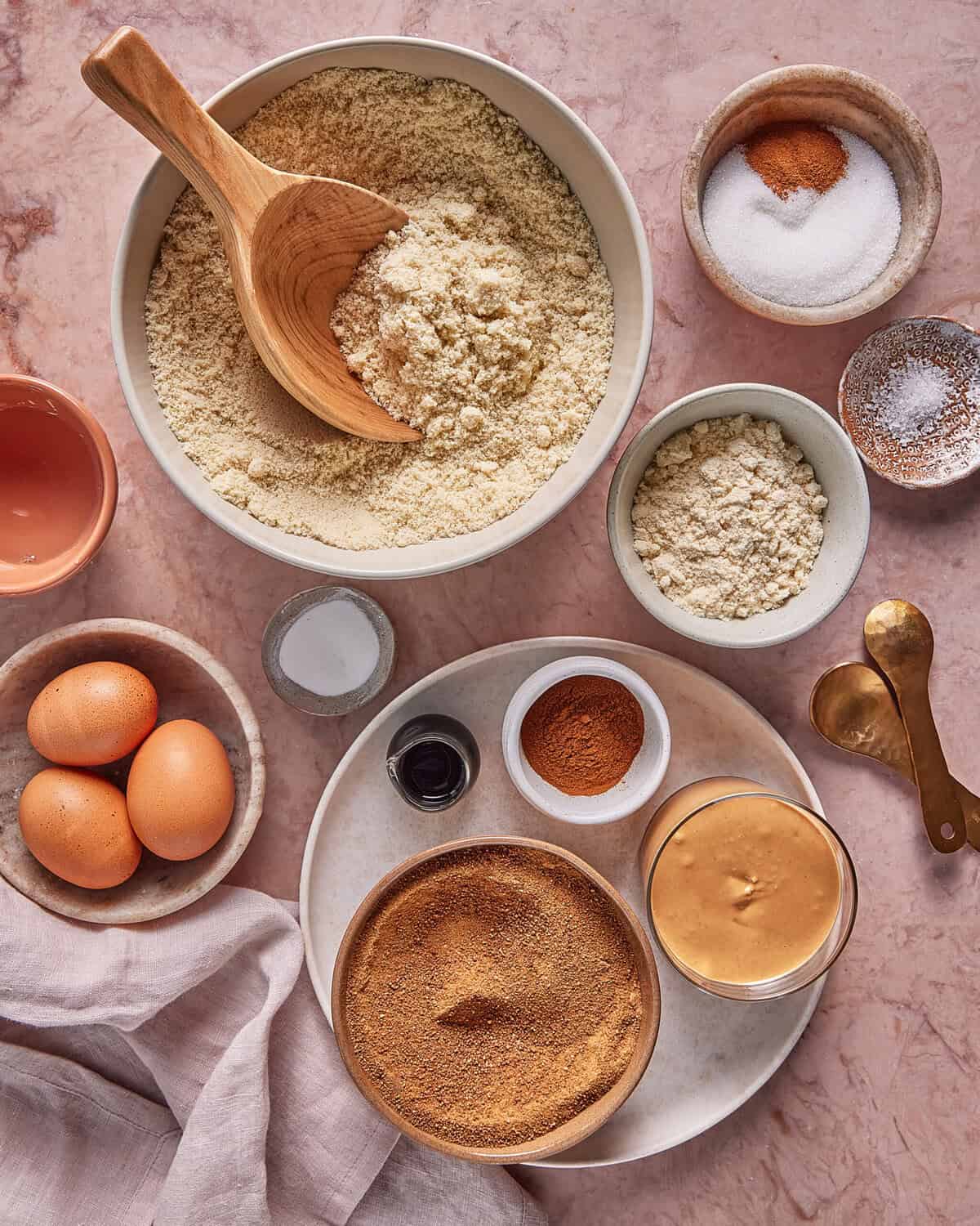 Top-down view of baking ingredients on a pink surface, including almond flour, sweetener, cinnamon, eggs, peanut butter, vanilla, salt, baking powder, and small measuring spoons. A wooden spoon rests in the flour.