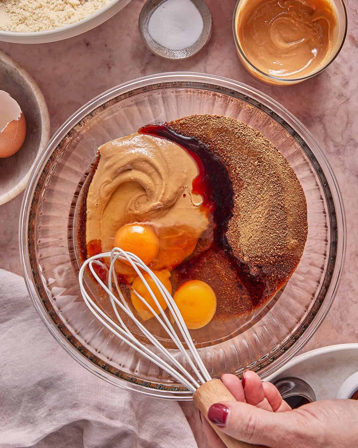 A hand holds a whisk over a bowl containing eggs, peanut butter, vanilla, and brown sugar, ready to be mixed. Nearby are a cracked egg, almond flour, salt, and more peanut butter on a pink surface.