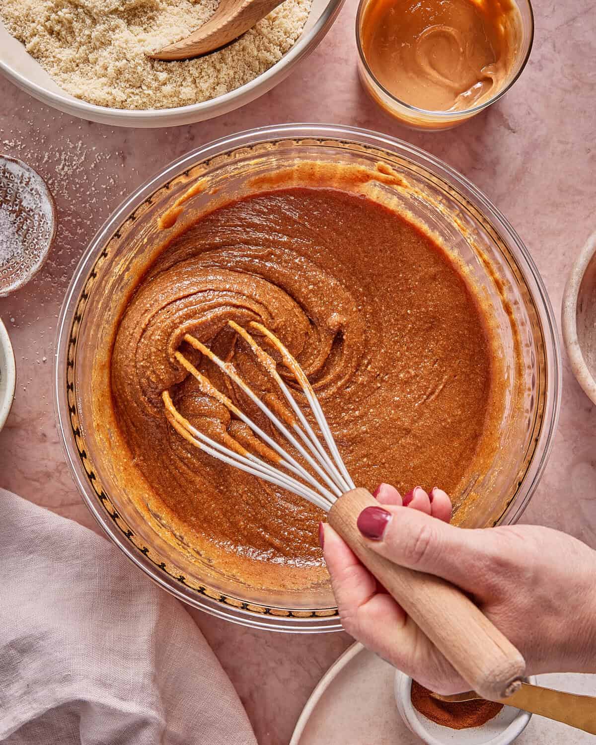 A hand holding a whisk mixes a thick brown batter in a glass bowl, surrounded by bowls of almond flour, peanut butter, and baking ingredients on a marble countertop.