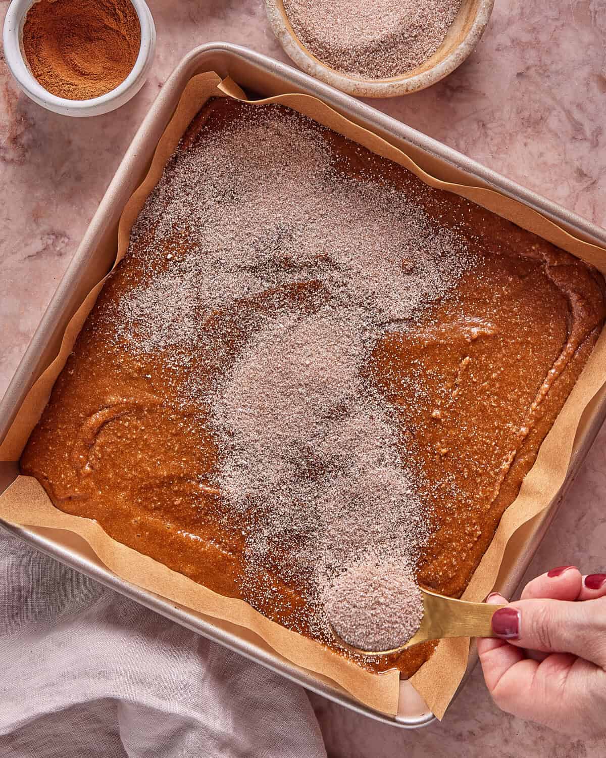 A hand sprinkles cinnamon sugar over unbaked batter in a parchment-lined square pan. Nearby are bowls containing cinnamon and cinnamon sugar, all on a light marble surface.