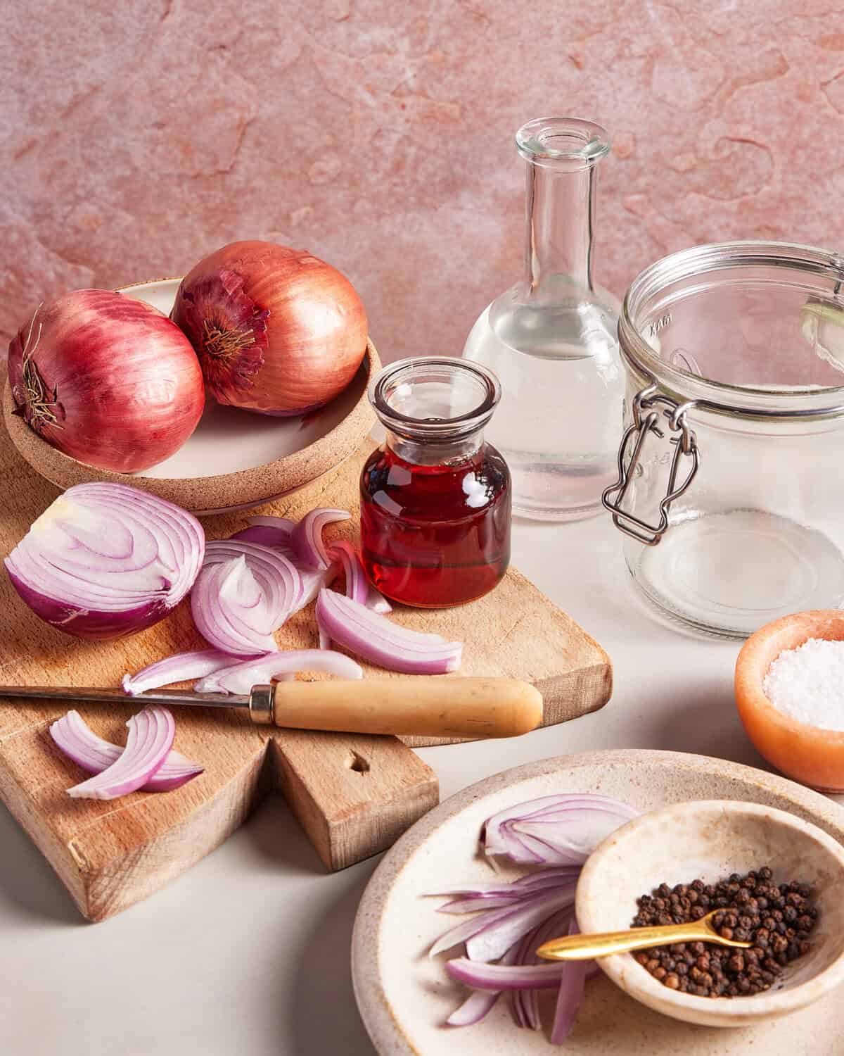 A kitchen scene with red onions, a glass bottle of vinegar, a glass bottle of water, a jar, salt, peppercorns, a knife, and sliced onions on a wooden cutting board against a pink background.