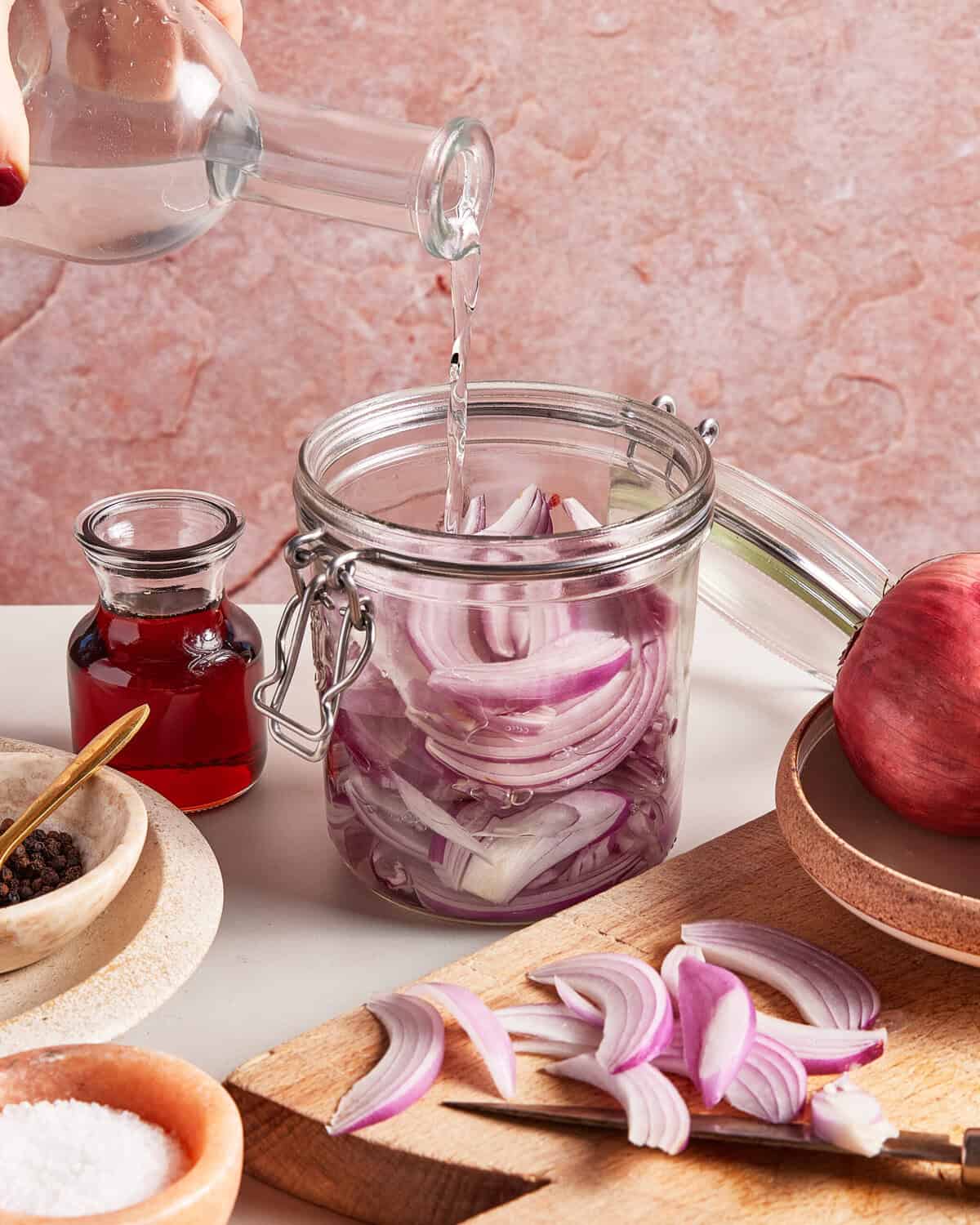 A hand pours vinegar into a glass jar filled with sliced red onions. Nearby are sliced onions on a cutting board, a small bottle of red vinegar, a bowl of salt, peppercorns, and a whole red onion.