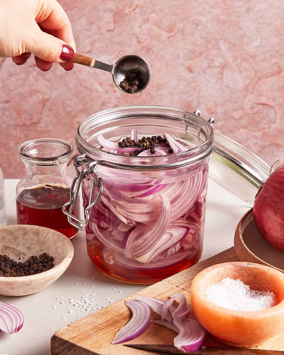 A hand sprinkles peppercorns into a glass jar filled with sliced red onions and red liquid. Nearby are small bowls of salt and pepper, a glass bottle of vinegar, and more onion slices on a cutting board.