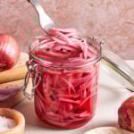 A hand uses a fork to lift pickled red onions from a glass jar filled with pink brine and peppercorns. Fresh red onions, salt, and peppercorns are arranged around the jar on a light surface.