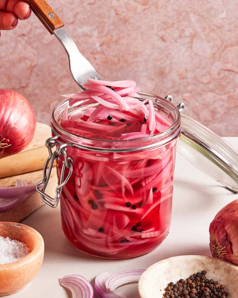 A hand uses a fork to lift pickled red onions from a glass jar filled with pink brine and peppercorns. Fresh red onions, salt, and peppercorns are arranged around the jar on a light surface.