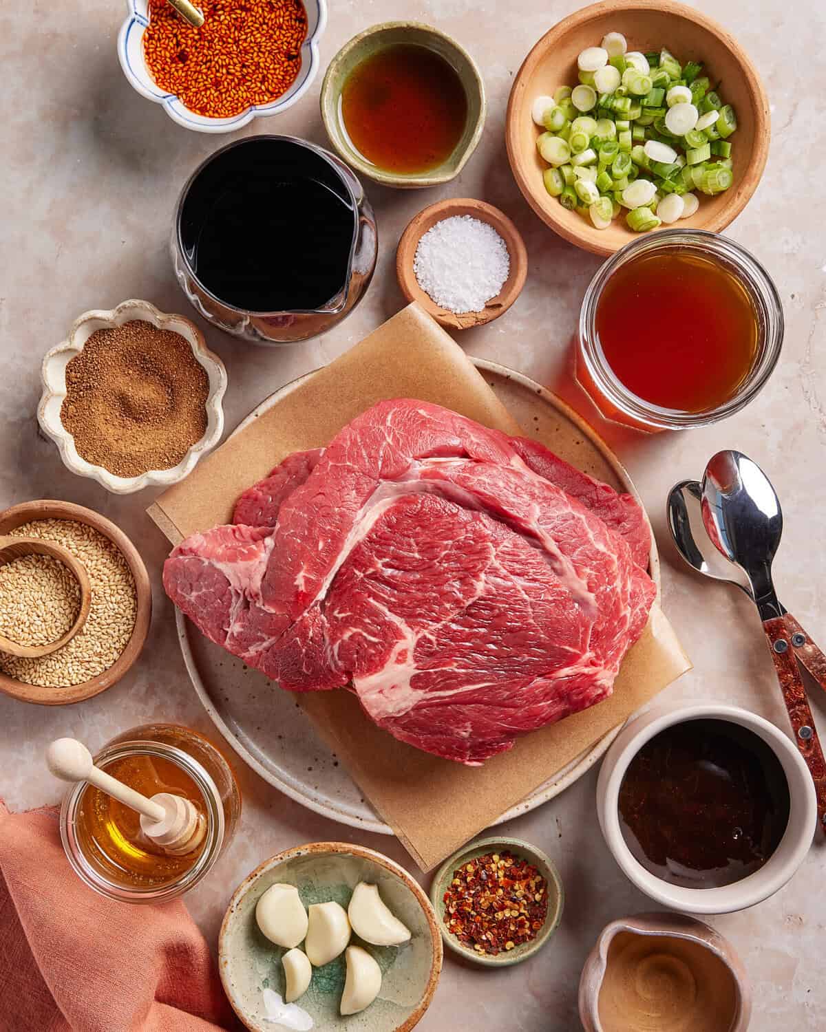 Overhead view of raw beef steak on parchment surrounded by bowls of sauces, seasonings, chopped green onions, garlic cloves, honey, sesame seeds, broth, and a spoon on a light countertop.