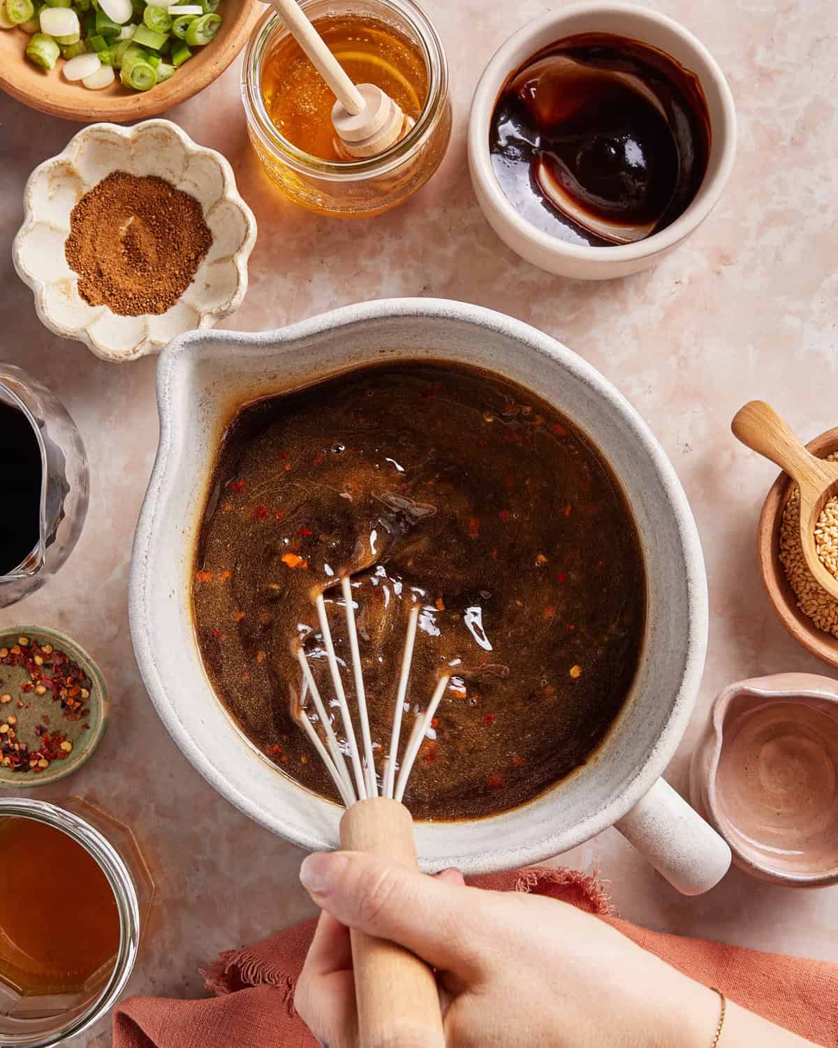 A hand whisks a dark sauce in a white bowl surrounded by small bowls of honey, hoisin sauce, spices, green onions, sesame seeds, and other ingredients on a light countertop.