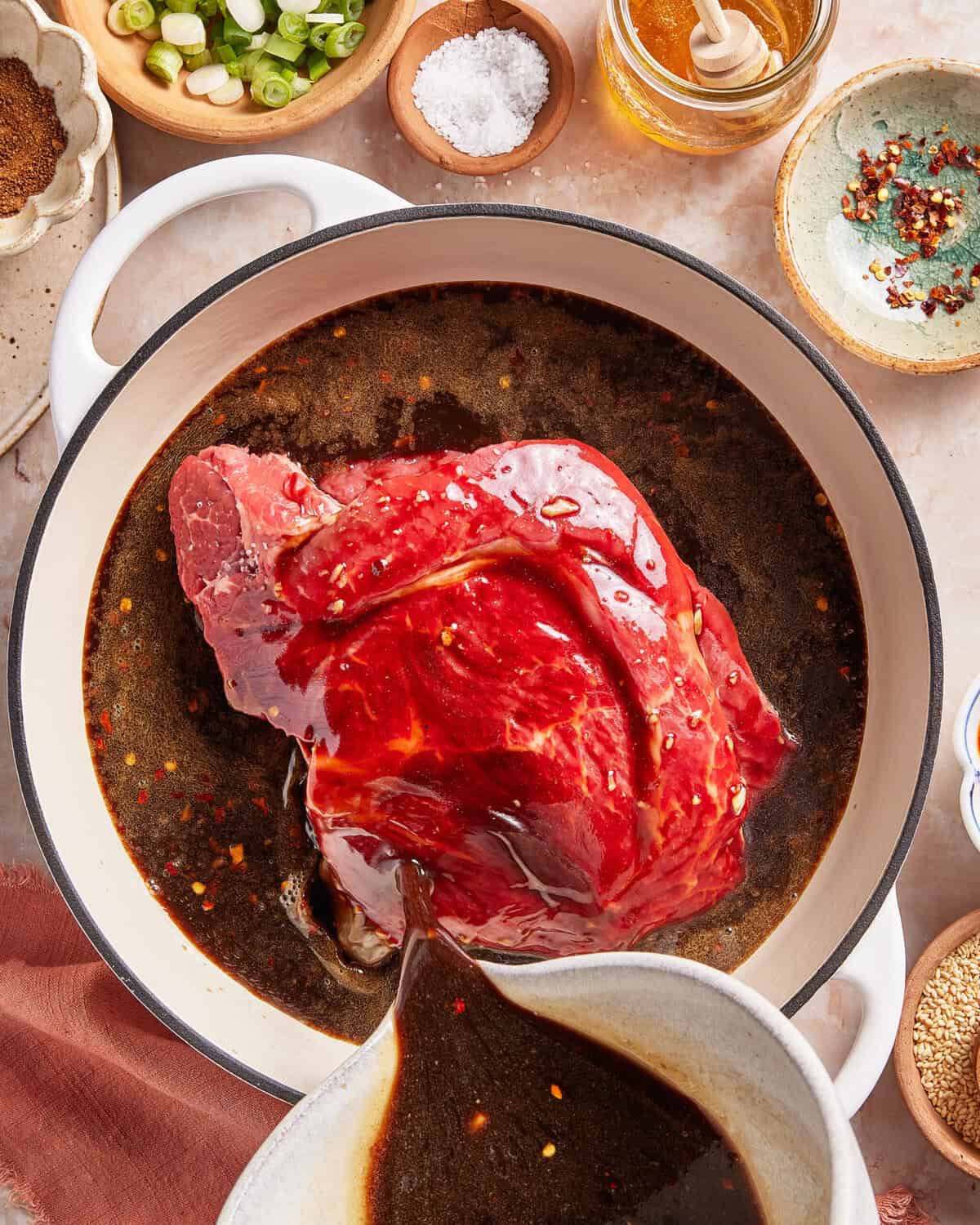 A raw beef roast sits in a pot of dark marinade, with more sauce being poured over it. Surrounding the pot are bowls of chopped green onions, salt, honey, red pepper flakes, and spices.