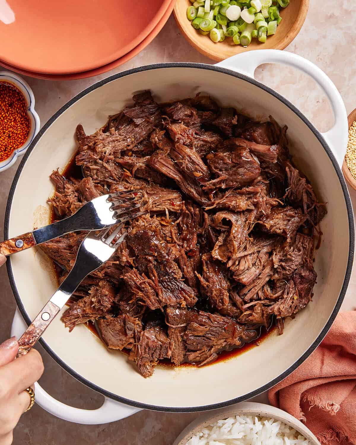 A hand uses two forks to shred tender, cooked beef in a white pot. Surrounding the pot are bowls of sliced green onions, rice, red spices, and empty plates on a marble surface.