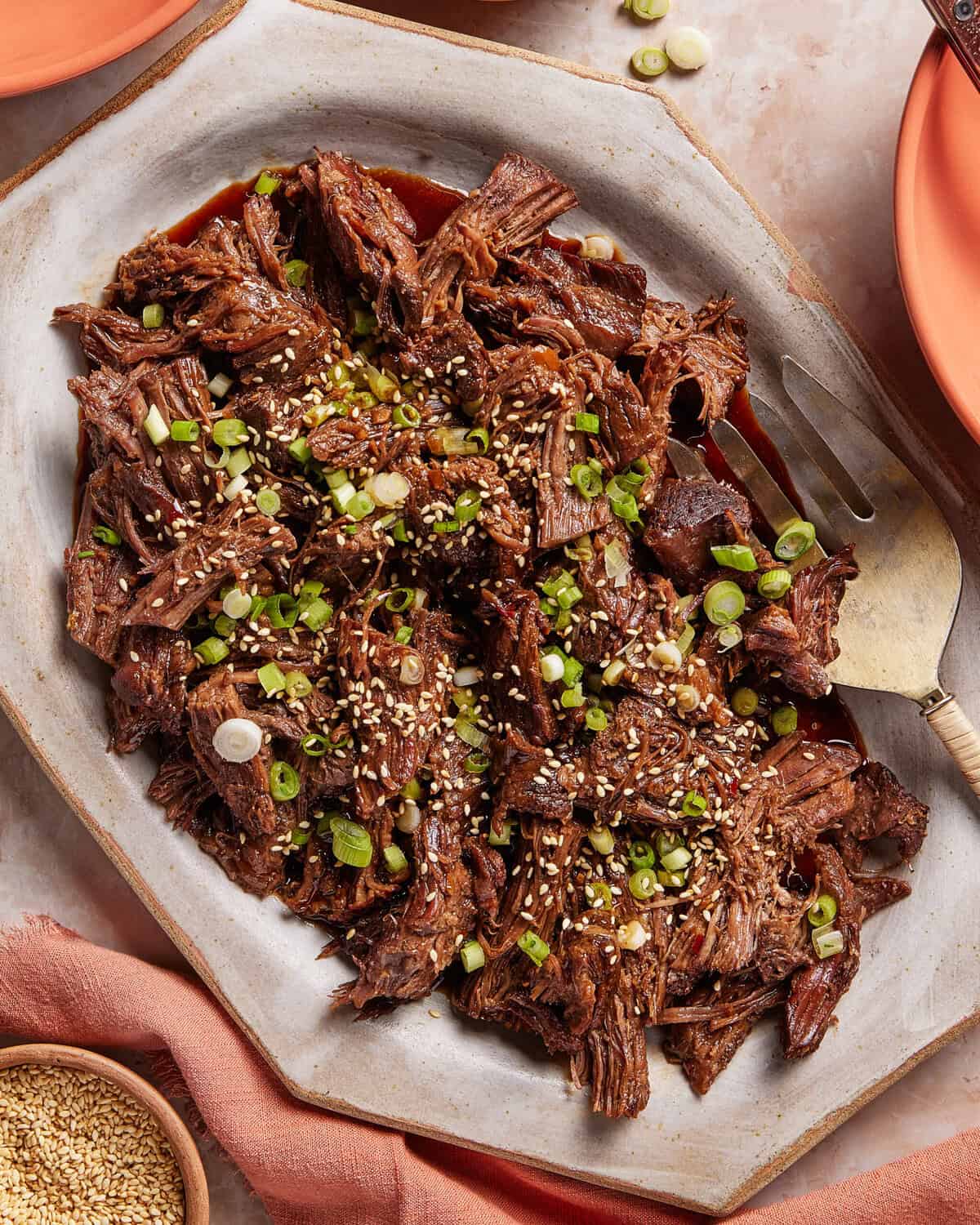 Shredded beef garnished with chopped green onions and sesame seeds is served on a white platter with a serving fork, surrounded by plates and a napkin.