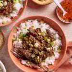 A bowl of white rice topped with shredded beef, sliced green onions, sesame seeds, and sauce, with a fork on the side. Additional bowls with rice, green onions, sesame seeds, and sauce are nearby.