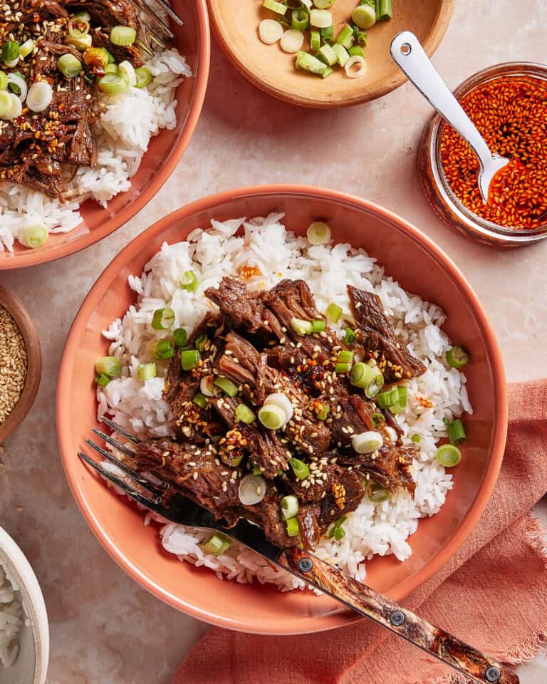 A bowl of white rice topped with shredded beef, sliced green onions, sesame seeds, and sauce, with a fork on the side. Additional bowls with rice, green onions, sesame seeds, and sauce are nearby.
