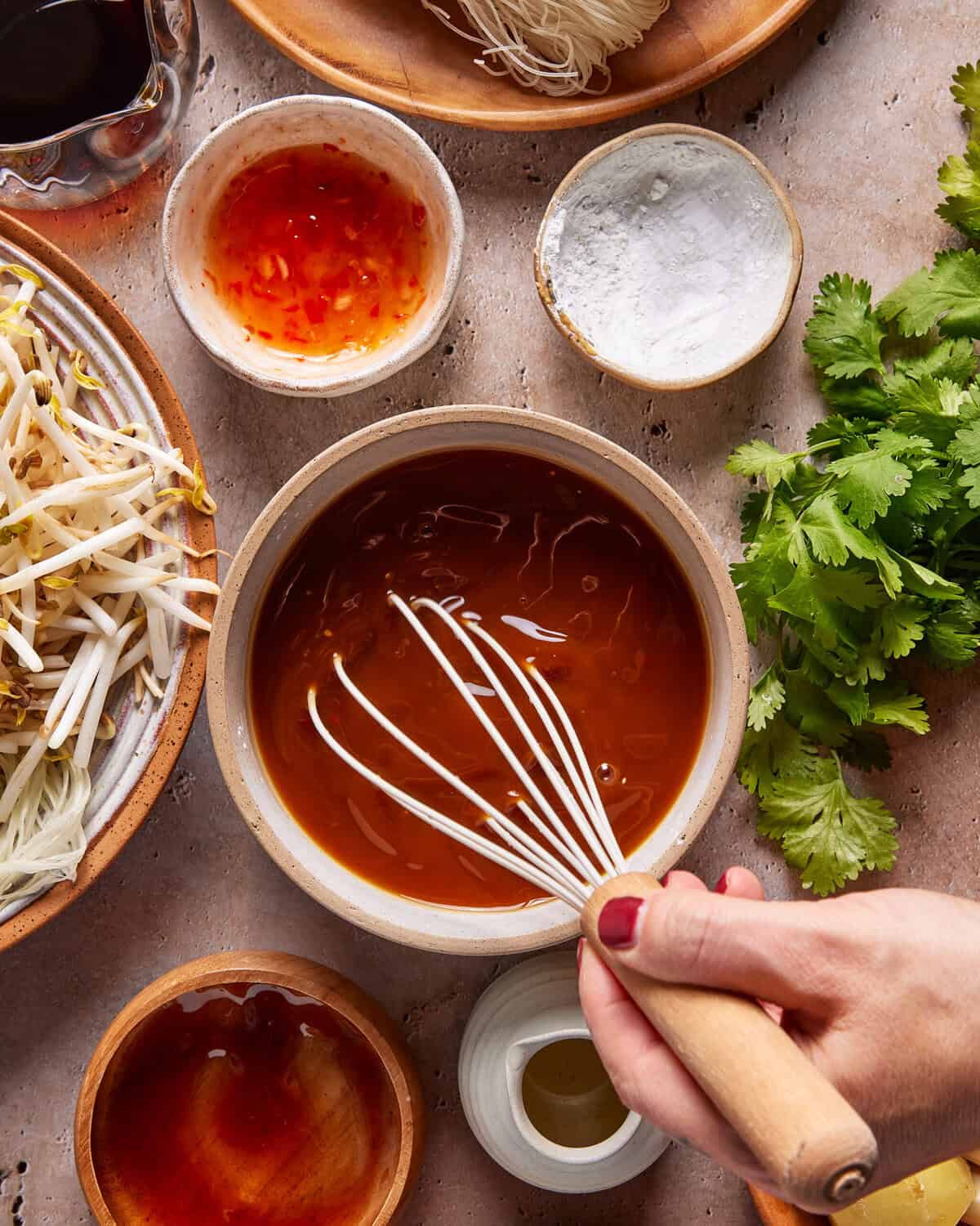 A hand using a whisk to mix sauce in a bowl, surrounded by fresh cilantro, bean sprouts, noodles, small bowls of sauce, and cornstarch on a light countertop.
