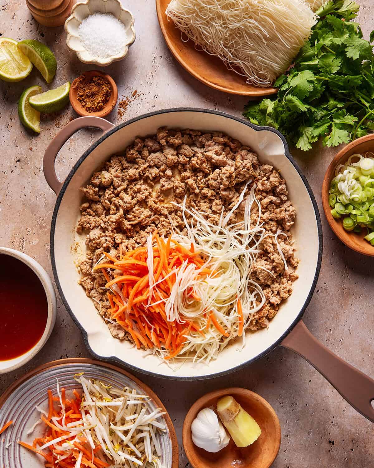 A skillet with cooked ground meat topped with shredded carrots and cabbage, surrounded by bowls of noodles, cilantro, green onions, bean sprouts, lime wedges, garlic, ginger, sauce, and spices on a rustic surface.