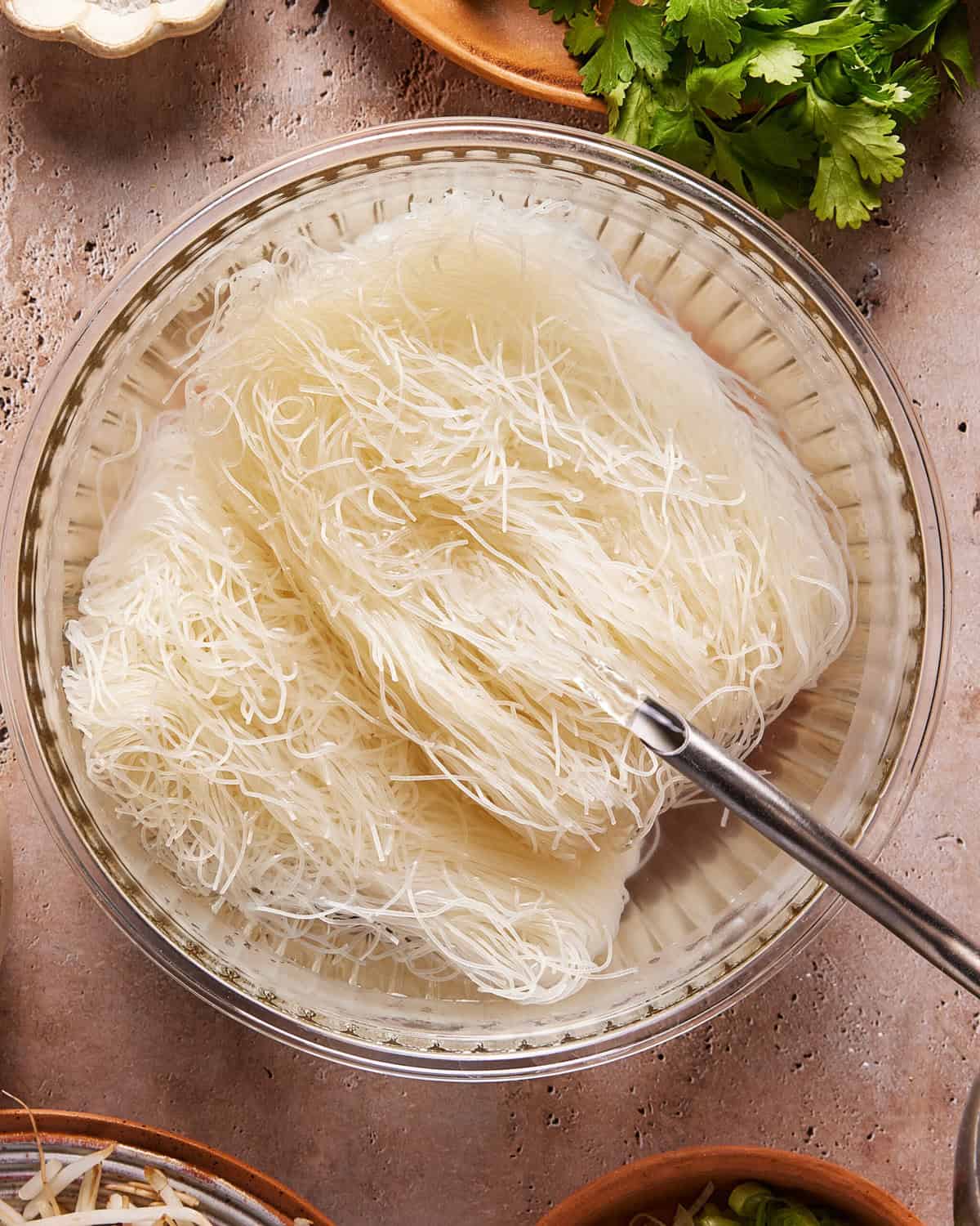 A glass bowl filled with soaked rice vermicelli noodles, with a metal fork resting inside. Fresh herbs and other ingredients are visible around the bowl on a textured surface.