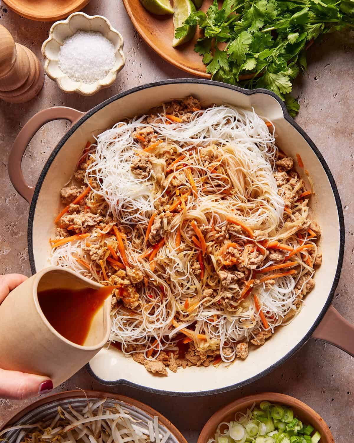 A hand pours sauce onto a skillet filled with rice noodles, shredded chicken, and julienned carrots. Surrounding the skillet are bowls of salt, lime, cilantro, bean sprouts, and sliced green onions.