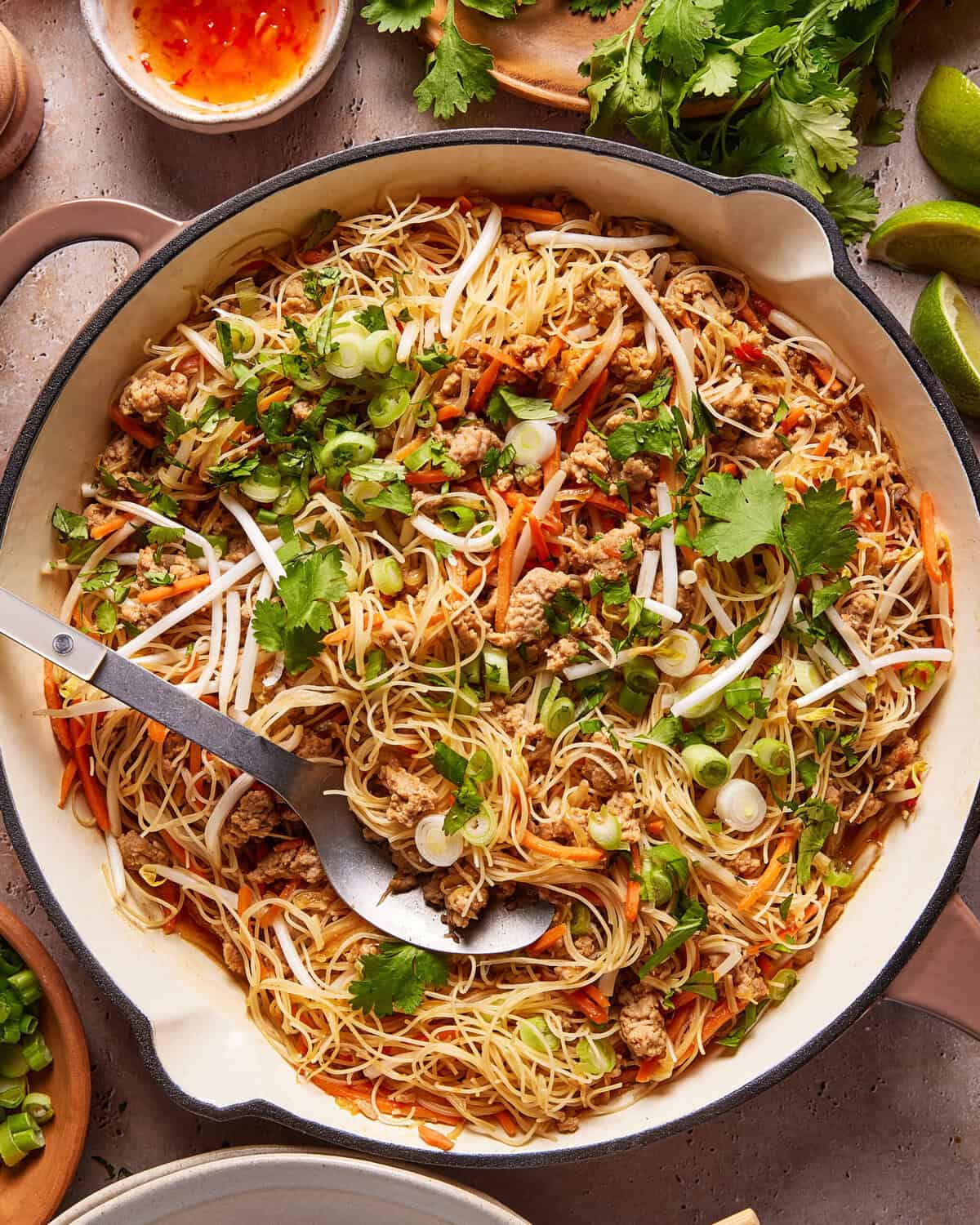 A large skillet filled with stir-fried rice noodles, ground meat, bean sprouts, shredded carrots, and green onions, garnished with fresh cilantro, with a serving spoon resting in the pan.