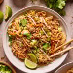 A bowl of stir-fried rice noodles with ground meat, shredded carrots, bean sprouts, green onions, and cilantro, served with lime wedges and chopsticks. Surrounding the bowl are herbs, sauce, and lime pieces.