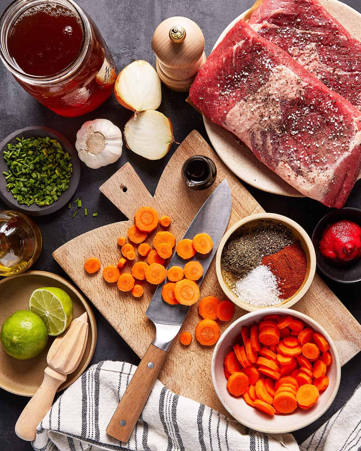Overhead view of raw beef on a plate, sliced carrots, a knife on a cutting board, bowls of chopped herbs and spices, garlic, onion, lime halves, tomato paste, olive oil, and a jar of broth on a dark surface.