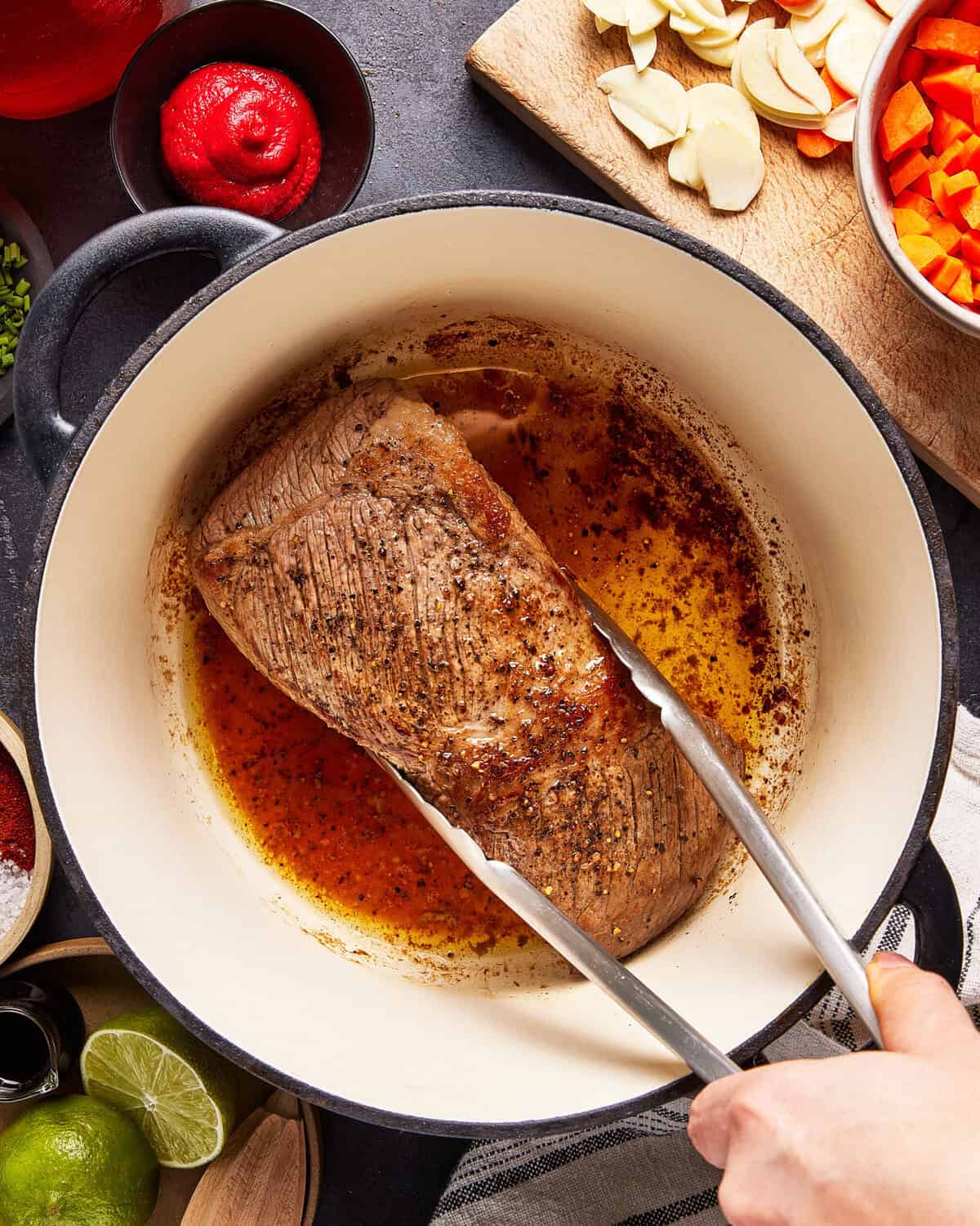 A hand uses tongs to sear a seasoned beef roast in a large white Dutch oven. Surrounding the pot are bowls of tomato paste, spices, and a cutting board with sliced carrots and garlic.
