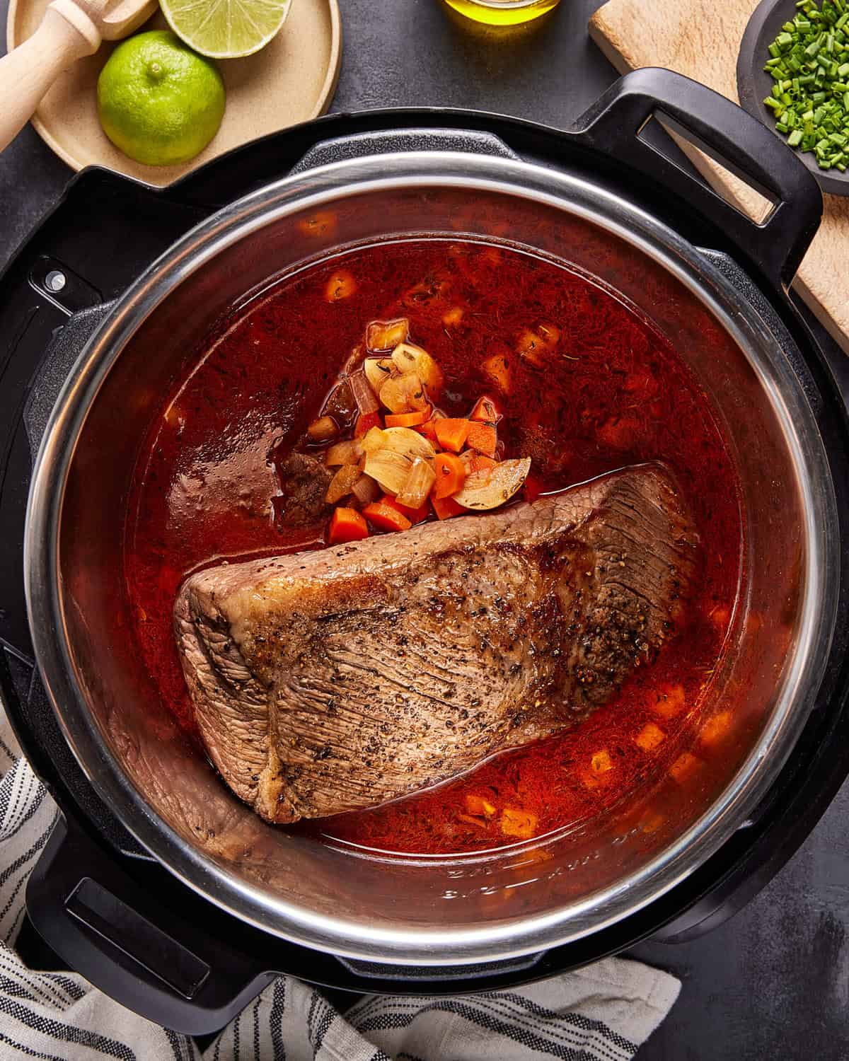 A large piece of seasoned beef brisket cooking in a red broth with chopped vegetables inside an Instant Pot, surrounded by a towel, lime, cilantro, and a wooden board.