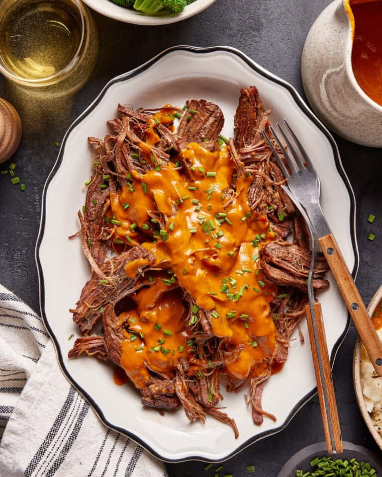 Shredded roast beef topped with orange sauce and chopped chives is served on a white plate with a fork and knife, next to a striped napkin, a drink, and a gravy boat.