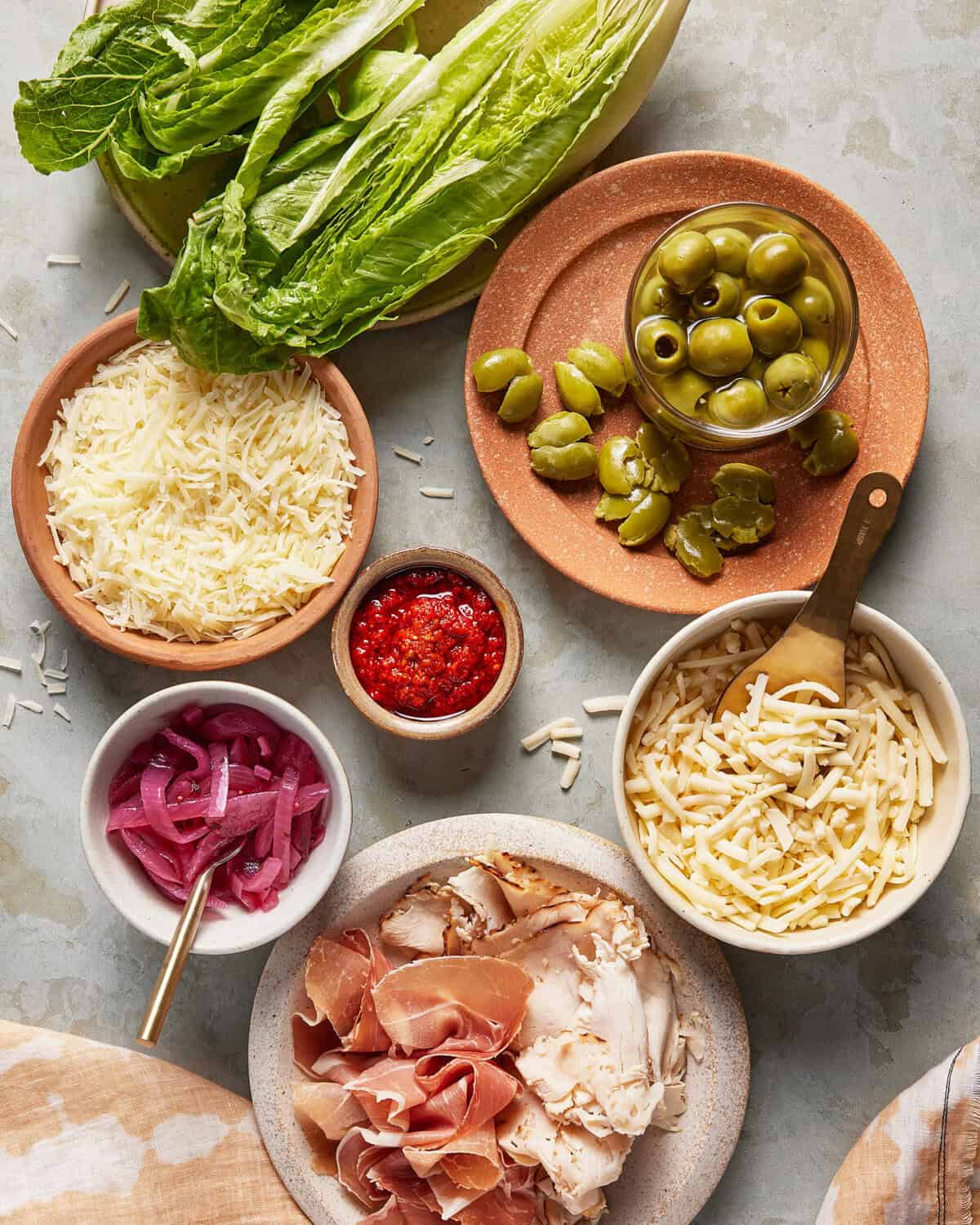 A top-down view of plates and bowls containing shredded cheese, green olives, red pepper spread, pickled red onions, shredded chicken, sliced prosciutto, and romaine lettuce leaves on a light surface.