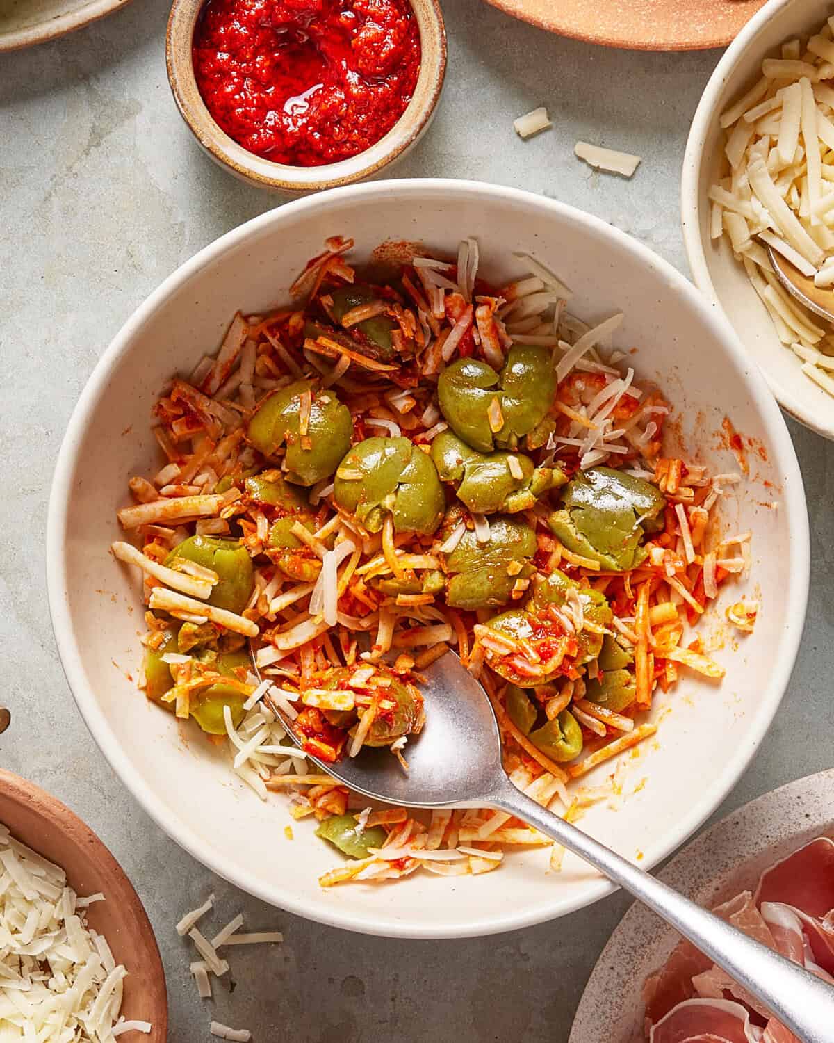 A bowl of shredded cheese, sliced green olives, and tomato sauce is being mixed with a spoon. Surrounding bowls hold more shredded cheese and red sauce.