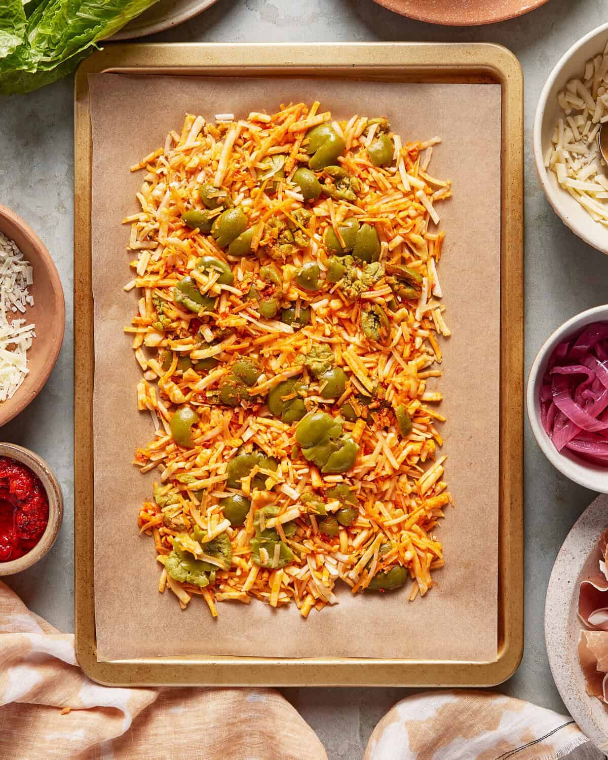 A baking tray lined with parchment paper holds a mixture of shredded cheese, chopped green peppers, and tomato sauce. Surrounding the tray are bowls containing shredded cheese, pickled onions, tomato paste, and greens.